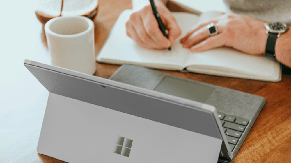 man writing in notebook infront of surface pro